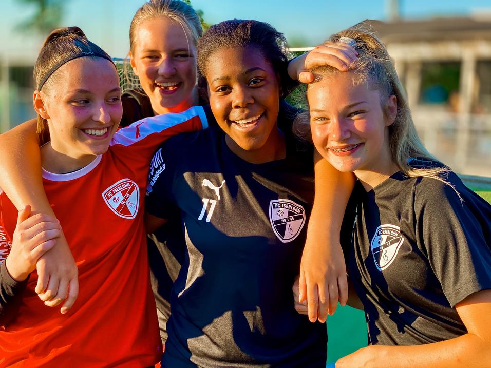 Vier junge Fußballspielerinnen in Teamtrikots posieren lachend gemeinsam auf dem Spielfeld.