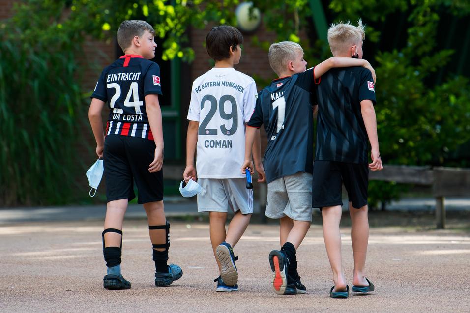 Vier Kinder in Fußballtrikots gehen Arm in Arm über einen Platz, umgeben von Bäumen und einer ruhigen Atmosphäre.