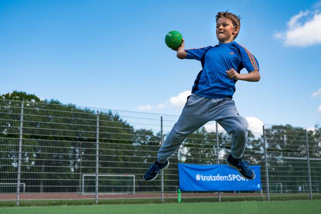 Junge springt auf einem Sportplatz und wirft einen grünen Handball, umgeben von einem Zaun und Bäumen im Hintergrund.