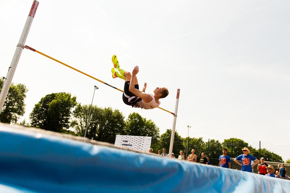 Springer beim Hochsprung über eine gelbe Lattenstange, im Hintergrund Zuschauer und grünes Umfeld.