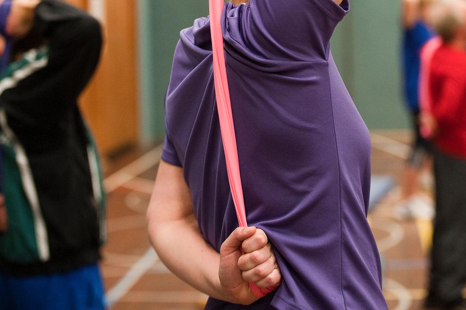 Frau in lila T-Shirt dehnt mit einem pinken Widerstandsband ihren Arm in einer Sporthalle. Hintergrund mit anderen Personen.